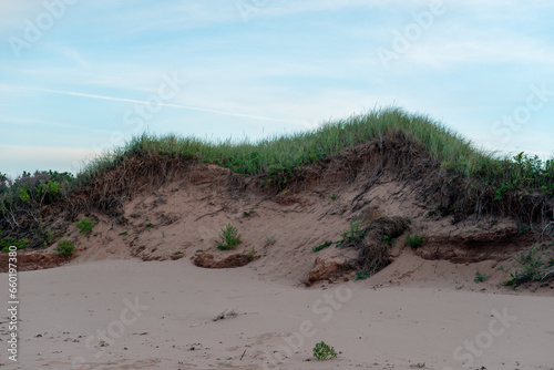Fototapeta Naklejka Na Ścianę i Meble -  A beach sand dune with a top layer of lush green grass. The windward mound of beige sand is eroding along the seashore in Prince Edward Island. The beach coastline is hills and valleys of fine grains.