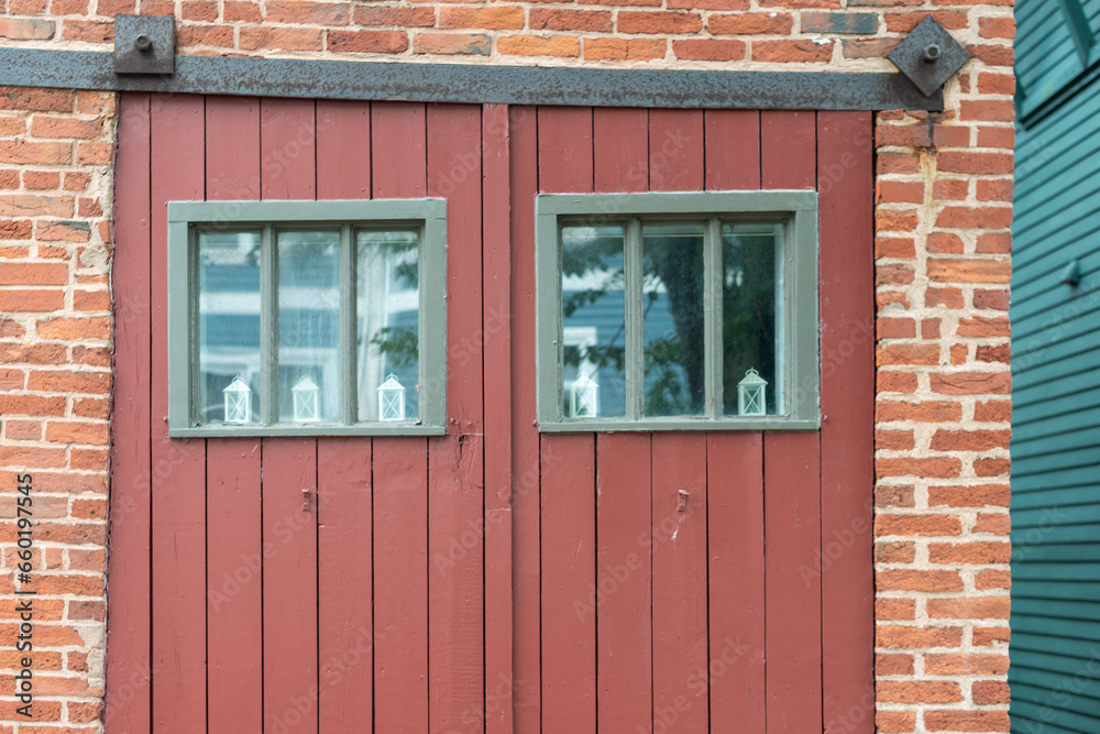 Closed double red wooden doors with small square glass windows. There's green wooden trim on the windows. The old barn doors are on metal rollers. The exterior building is red brick with white mortar. Closed double red wooden doors with small square glass windows. There's green wooden trim on the windows. The old barn doors are on metal rollers. The exterior building is red brick with white mortar.