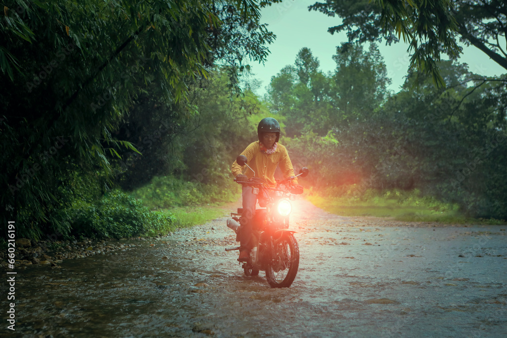 man riding small endurom motorcycle crossing shallow creek among rain falling at forest - obrazy, fototapety, plakaty 