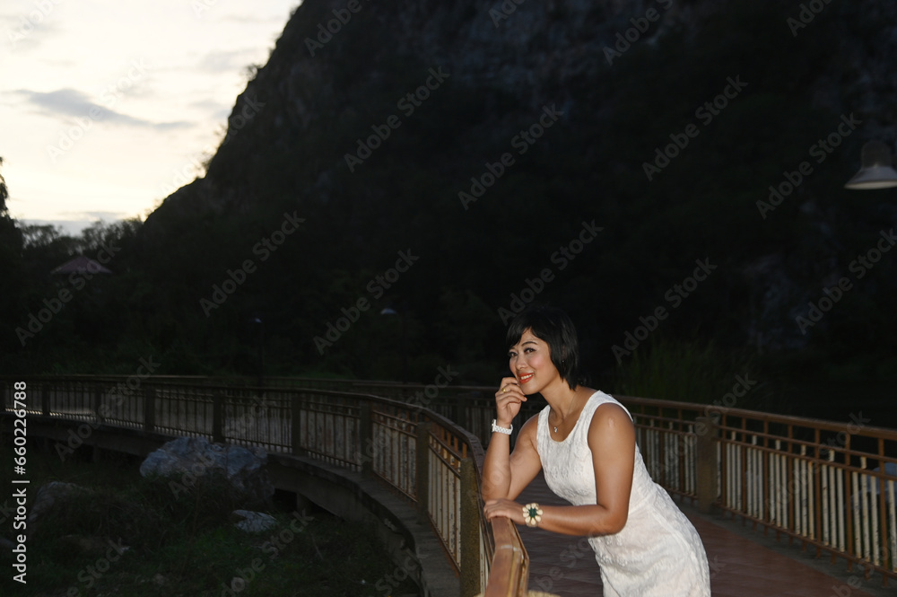 Portrait photography of a mature, beautiful, happy Asian woman. Model in white casual clothes. Standing and smiling at the camera, standing on a bridge with mountains behind.