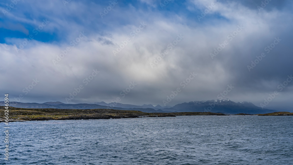 Landscape of the Beagle Canal. The islet and the stunted vegetation of Patagonia are visible in the distance. Ripples on the blue water. Andes mountain range against the sky and clouds. Argentina. 