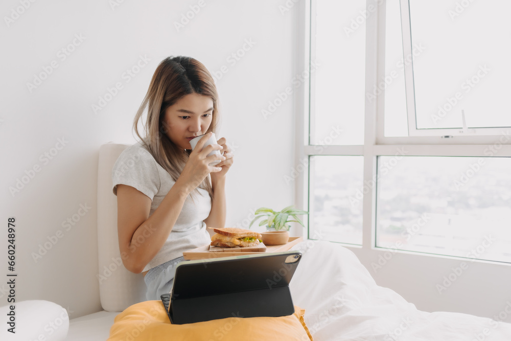 Asian Thai woman having breakfast sandwich and drinking hot coffee on white bed while looking or watching tablet, working at home alone on chilling day. Asian Thai woman having breakfast sandwich and drinking hot coffee on white bed while looking or watching tablet, working at home alone on chilling day.