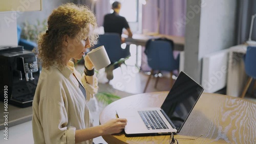 Pretty curly woman enjoying fresh coffee on workplace while types on modern laptop keyboard against a blurry background with colleague in light company office, side view, medium shot