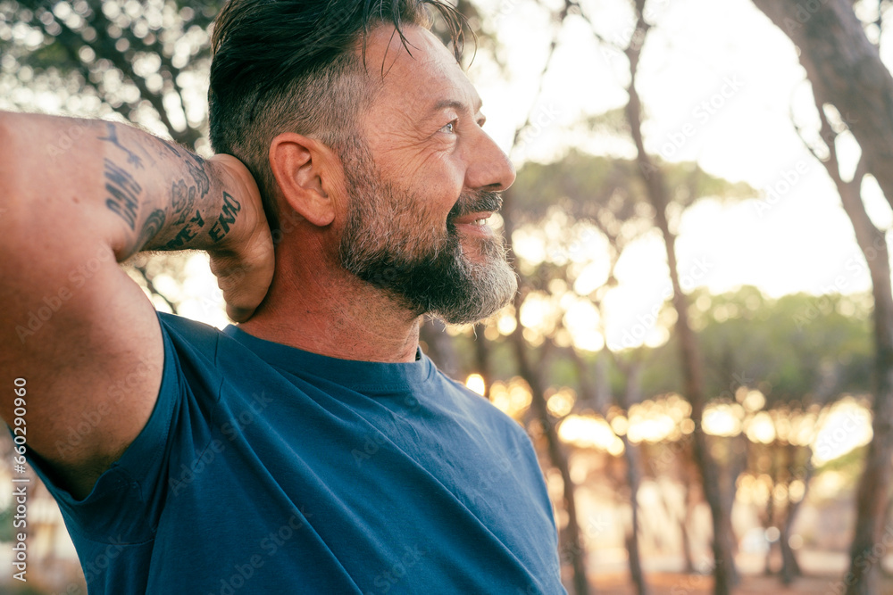 Side view of happy adult man enjoying leisure outdoor activity at the park touching his head and smiling with trees and sunset light in background. People and nature connection. Serene person outside - obrazy, fototapety, plakaty 