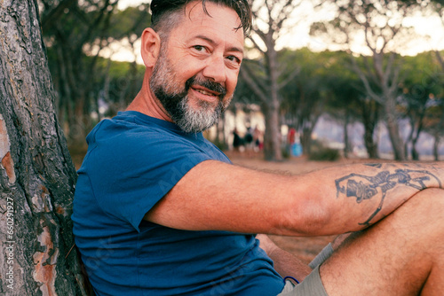 Fototapeta Naklejka Na Ścianę i Meble -  Side closeup portrait of adult man with beard smiling and enjoying relax time at the park. Happy and serene people with cheerful expression. Looking on camera. Handsome male 50 years old outside.