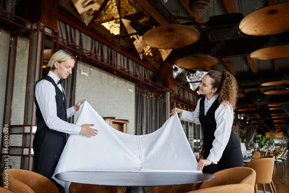 Side view portrait of two elegant servers covering tables with white tablecloth while preparing luxury restaurant for opening, copy space