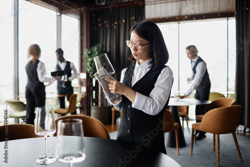 Side view portrait of Asian young woman as waiter polishing glasses in luxury restaurant, copy space