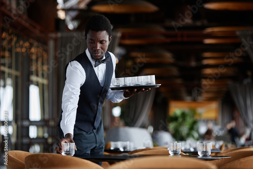 Waist up portrait of elegant Black man as waiter setting tables with crystal glasses preparing for restaurant opening, copy space