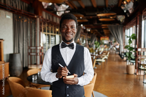 Waist up portrait of young Black man as server in luxury restaurant smiling at camera and holding notepad ready to take orders, copy space