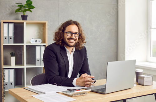 Happy businessman, auditor or financial accountant in office. Cheerful long haired young business man in suit and glasses sitting at work desk with laptop and calculator, looking at camera and smiling