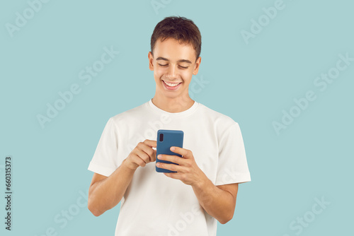 Happy teenager using modern cell phone. Studio portrait adolescent boy in T shirt standing isolated on light blue background holding smartphone, scrolling news feed, looking at funny memes and smiling © Studio Romantic