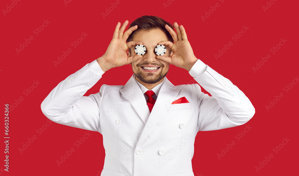 Portrait of happy lucky confident handsome man in elegant white suit standing on red studio background, smiling and covering both his eyes with two poker chips in hands. Casino, gambling, luck concept