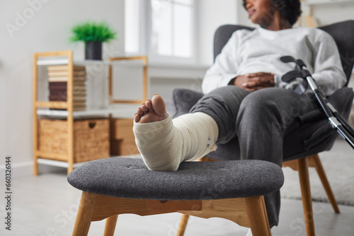 Cropped view of woman with leg in plaster bandage and crutch sittingin armchair at home. Selective focus of sitting African American woman with broken leg. Treatment at home, recovery concept