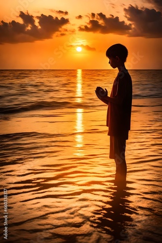 The silhouette of a young boy stands in a posture of prayer and adoration against the backdrop of a breathtaking sunset over the sea. The golden hues of the sun's descent cast a warm glow on the water photo