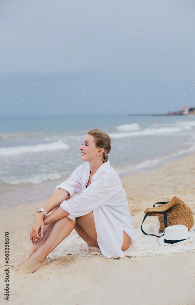 a young fair-skinned woman in a white shirt sits alone on the coast against the background of the sea