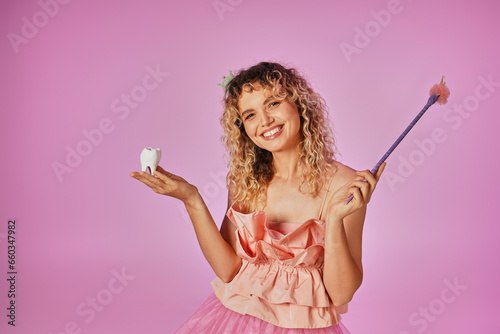 joyous curly haired tooth fairy in pink costume and headband holding baby tooth and magic wand photo