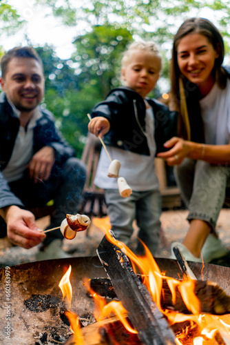 Close-up of a young family dad, mom and daughter sitting together by fire in the forest and roasting marshmallows near their country house