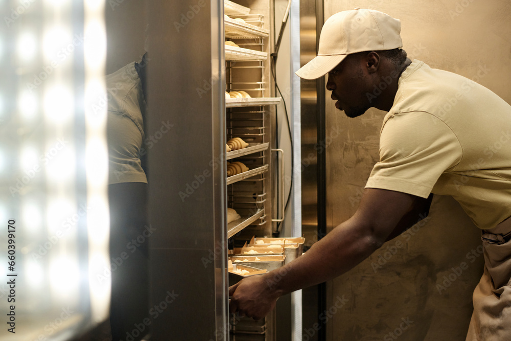 Side view portrait of young male baker putting fresh pastries in freezer at bakery kitchen, copy space