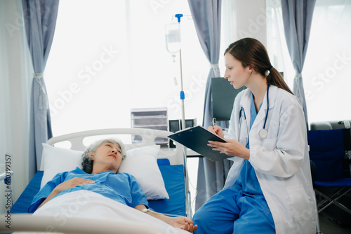 Doctor in white suit take notes while discussing and Asian elderly  woman patient who lying on bed with receiving saline solution in hospital or clinic.