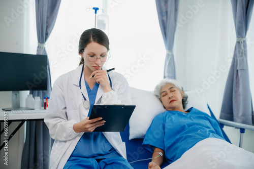 Fototapeta Naklejka Na Ścianę i Meble -  Doctor in white suit take notes while discussing and Asian elderly, woman patient who lying on bed with receiving saline solution in hospital or clinic.
