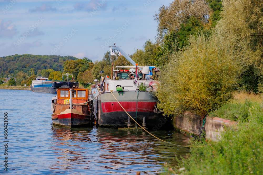 Two boats anchored at a dock berth on Albert Canal, cargo ship in background, abundant wild vegetation and mountains with green trees against misty blue sky, sunny summer day in Kanne, Riemst, Belgium