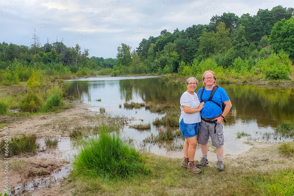 Senior adult couple of hikers standing at muddy bank of a stream among wild vegetation and leafy trees in background against blue sky, Dutch nature reserve Brunssummerheide, South Limburg, Netherlands