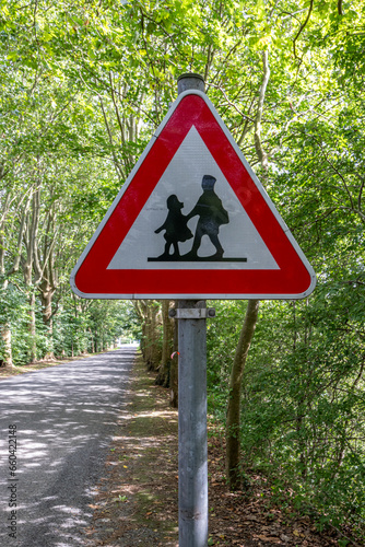 School warning sign on side of a rural road between huge green leafy trees, fading into background, drawing of two children walking in a red triangle, sunny summer day in Lanaye, Vise, Belgium