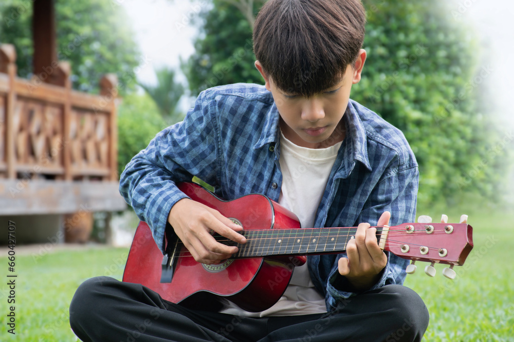 Obraz premium Asian cute boy playing guitar in the front yard of his own house happily, soft focus, concept for recreational activity and free times hobbies of children around the world.