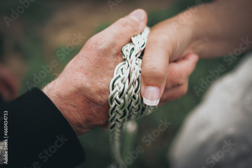 Close up picture of bride and groom's hands tied with white and green rope.