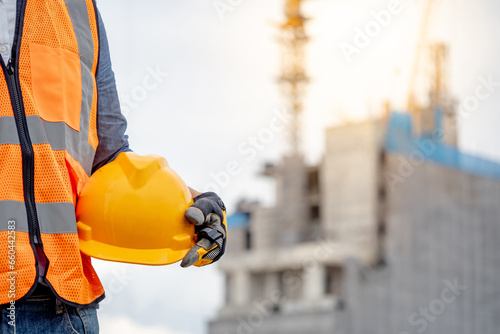 Safety workwear concept. Male hand holding yellow safety helmet or hard hat. Construction worker man with reflective orange vest and protective gloves standing at unfinished building with tower crane