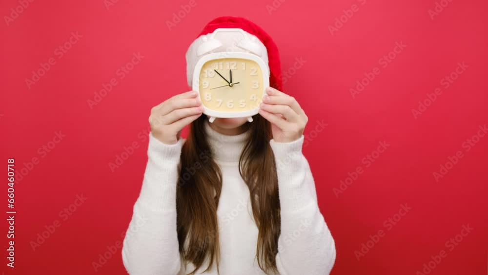 Cute young woman wears warm sweater and Santa hat hold in hand cover face with clock show five to twelve time, posing isolated on pastel red studio background wall. Happy New Year 2024 holiday concept