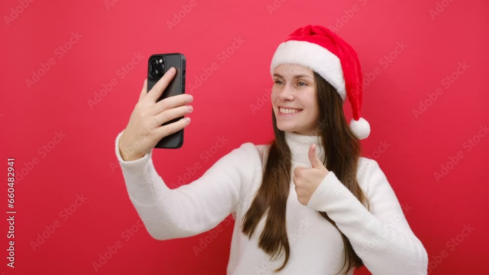 Cute young woman wear white knitted sweater and Santa hat posing doing selfie shot on mobile cell phone show thumb up, isolated on pastel red background in studio. Happy New Year 2024 holiday concept Cute young woman wear white knitted sweater and Santa hat posing doing selfie shot on mobile cell phone show thumb up, isolated on pastel red background in studio. Happy New Year 2024 holiday concept