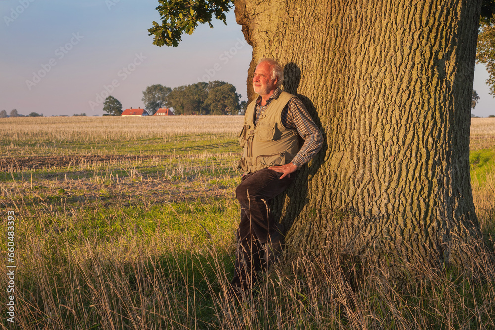 An elderly man leans against the mighty trunk of an oak tree, basking in the evening sun, savoring nature's tranquility. An elderly man leans against the mighty trunk of an oak tree, basking in the evening sun, savoring nature's tranquility.