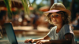 Little boy in straw hat is playing computer game or watching cartoon by laptop in a cafe on a tropical beach while food is cooking. Modern technology for education and entertainment. Vacation concept