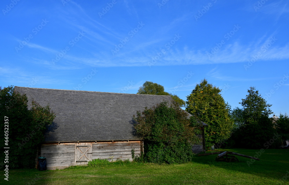 Obraz premium A view of an old wooden hut, shack, or stable surrounded with growing flowers, herbs, a wooden fence, and ancient farming equipment seen next to a well maintained lawn, pastureland, or meadow