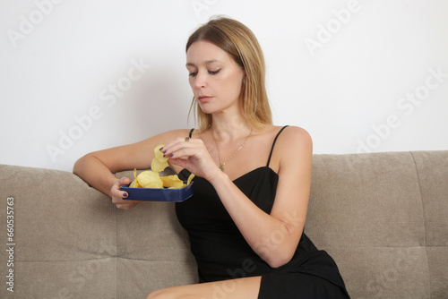 Young sad woman having unhealthy snack, eating potato chips in the livingroom. Junk food and unhealthy eating concept.
