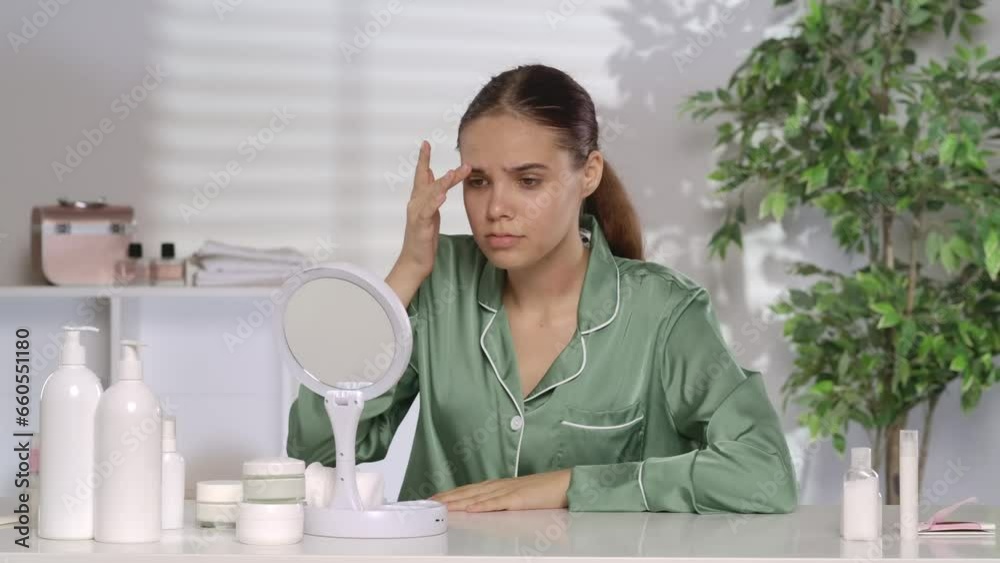 A young woman with problematic facial skin looks disappointedly in the mirror. A woman without makeup and wearing green pajamas sits at her dressing table in the morning. The concept of problem skin. A young woman with problematic facial skin looks disappointedly in the mirror. A woman without makeup and wearing green pajamas sits at her dressing table in the morning. The concept of problem skin.