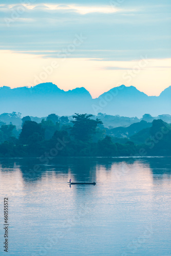 Silhouette of a fishing boat in the Mekong River, bordering Thailand and Laos, in the morning. The background is mountains. There is soft yellow sunlight shining, vertical image suitable for postcards