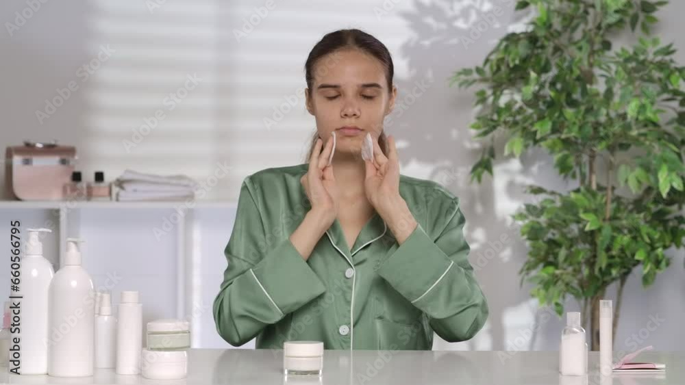 A young woman cleanses her face with cotton pads. A woman in green pajamas sits at a dressing table in front of a mirror, performing her morning routine. Slow motion. A young woman cleanses her face with cotton pads. A woman in green pajamas sits at a dressing table in front of a mirror, performing her morning routine. Slow motion.