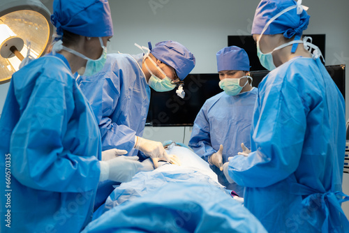 Side view of Caucasian male surgeon with his team, in blue surgical uniforms, face masks, and medical loupes, standing around the operating bed and performing surgery on a patient in an operating room