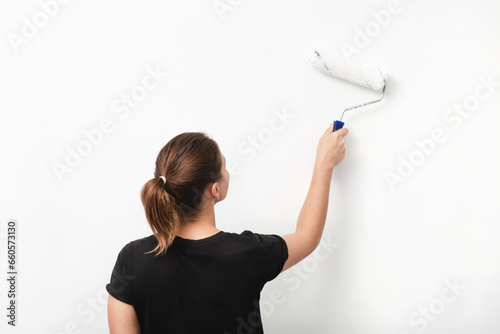 a young woman in a black T-shirt from the back paints a wall with a paint roller with white paint. mature woman on a white background in dark clothes with a paint roller paints a wall. medium shot.