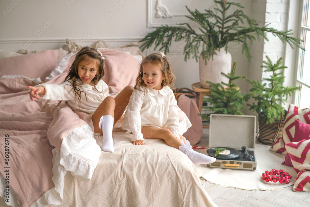 Children are sitting on a bed early on Xmas morning in a bedroom decorated with Christmas trees. A curious girl points her hand to the side Children are sitting on a bed early on Xmas morning in a bedroom decorated with Christmas trees. A curious girl points her hand to the side