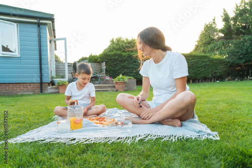 Canvas Print Happy Young mother and preschooler son having outdoor picnic dinner, eating pizza sitting on backyard Lawn on Sunny Day