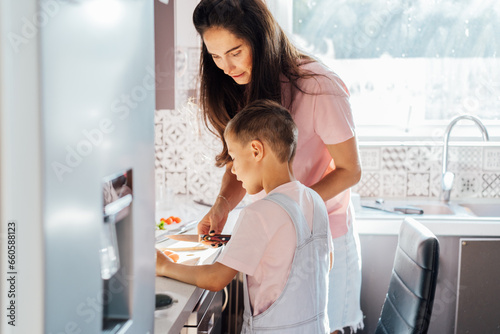 Loving young mother teaching her preschooler son prepare healthy food or salad at home kitchen  caring happy mom cooking together with kid on modern kitchen  boy involved in preparing food with mum