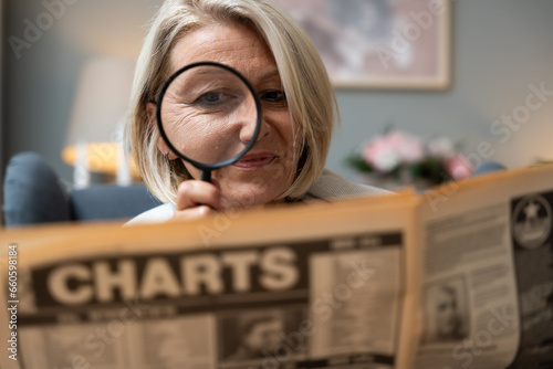 Senior woman reading newspaper with magnifying glass, unaware making funny facial expressions. Middle age retired female having fun photo