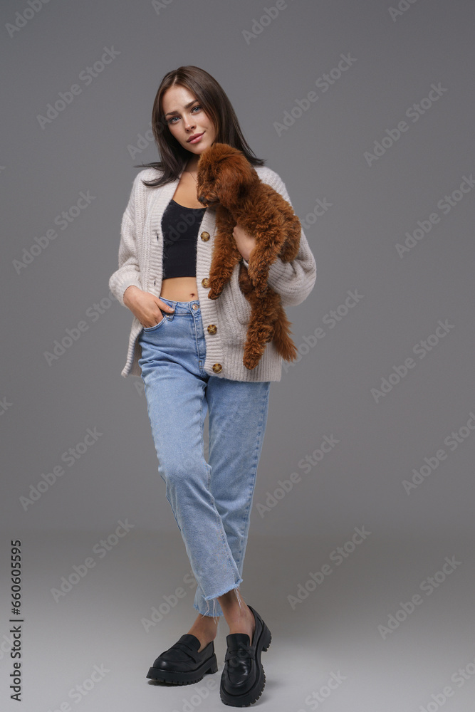 Stylish young brunette woman in casual wear cradling her cherished brown toy poodle, posed before a gray studio backdrop Stylish young brunette woman in casual wear cradling her cherished brown toy poodle, posed before a gray studio backdrop