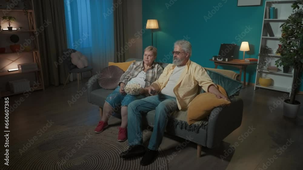 In the frame, an elderly couple are sitting on the couch and choose which movie or program to watch on TV and eat popcorn. They have chosen, watch with joy and talk about something In the frame, an elderly couple are sitting on the couch and choose which movie or program to watch on TV and eat popcorn. They have chosen, watch with joy and talk about something