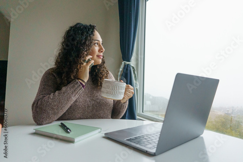 Curly hair young woman working on laptop in cozy nook with mug	