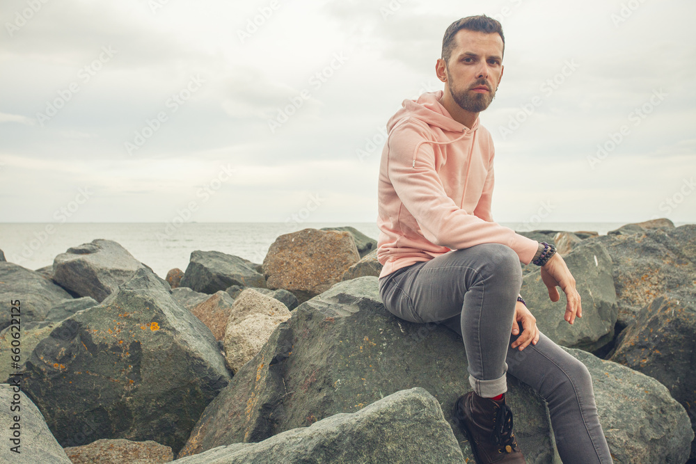 Street fashion concept. Young bearded man sitting in trendy clothing on the stony sea shore. Cloudy weather. Text space. Outdoor shot Street fashion concept. Young bearded man sitting in trendy clothing on the stony sea shore. Cloudy weather. Text space. Outdoor shot