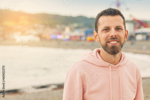 Welcome concept. Young bearded man wearing pink trendy hoodie and smiling, posing over sea coast on backgound. Cloudy weather. Text space. Outdoor shot